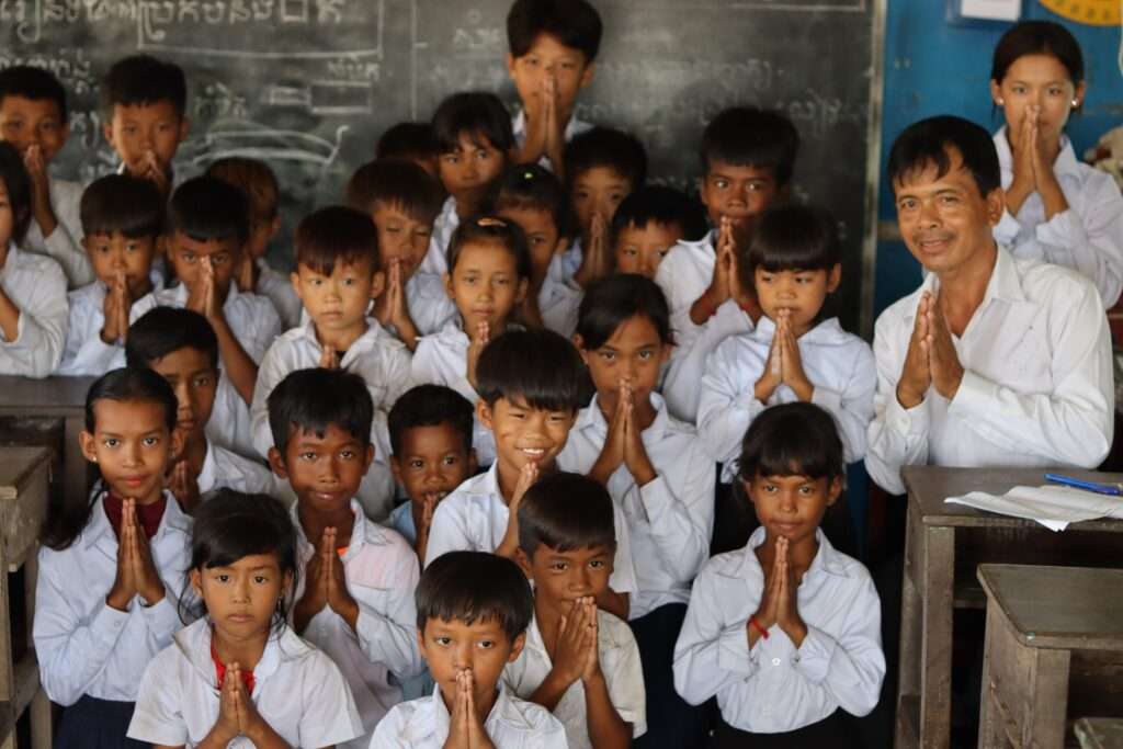 Cambodian schoolchildren and their teacher greeting visitors with traditional sampeah gesture in a classroom supported by ActionAid Cambodia.