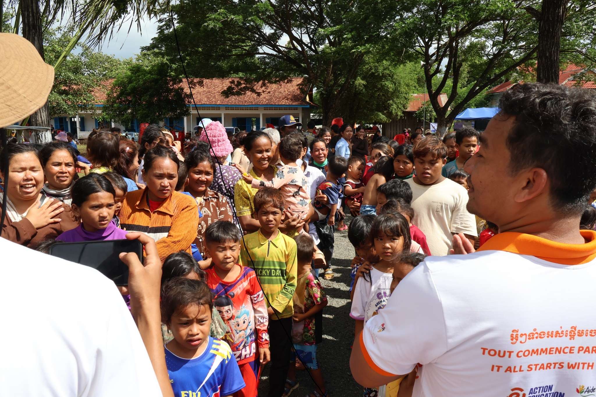 Aide et Action Cambodia team distributing education kits to families and children in a post-conflict zone, with a large crowd gathered at a school compound.