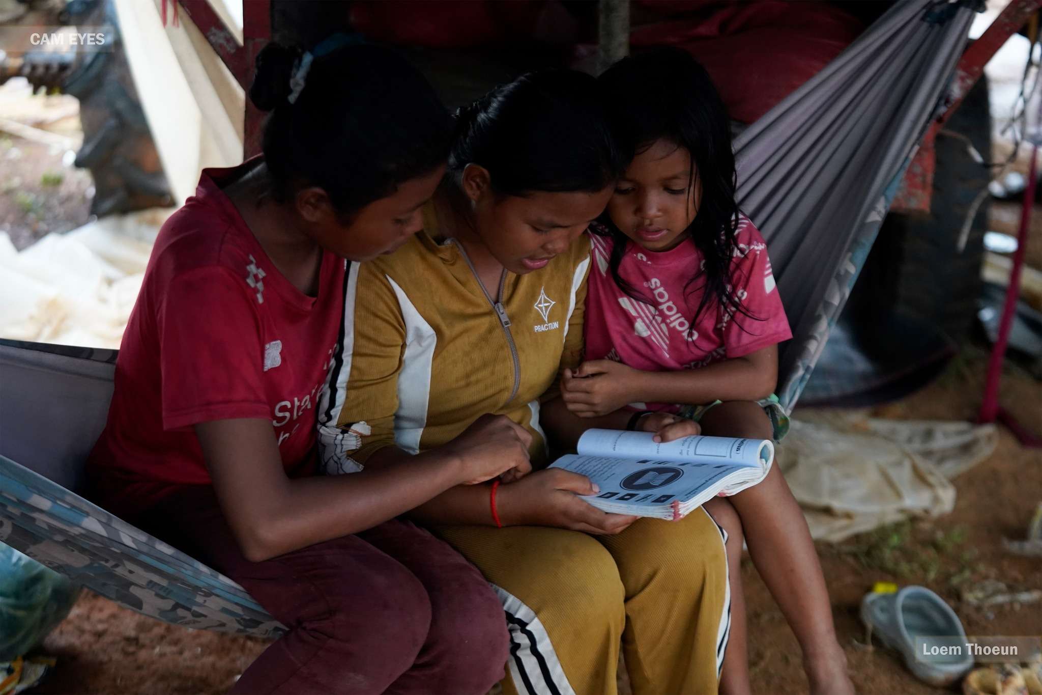 Three displaced Cambodian children read a book together in a temporary shelter, highlighting the importance of education during times of crisis.