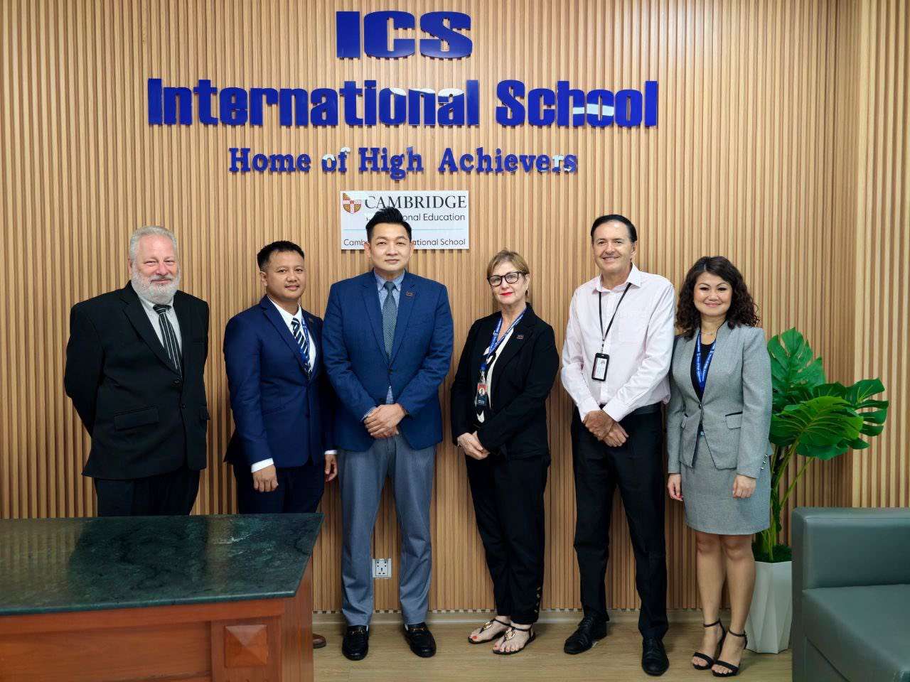 Group photograph of ICS International School leadership team and BritCham Cambodia representatives celebrating the school’s membership, taken inside the ICS campus lobby with the “Home of High Achieve-rs” banner in the background.