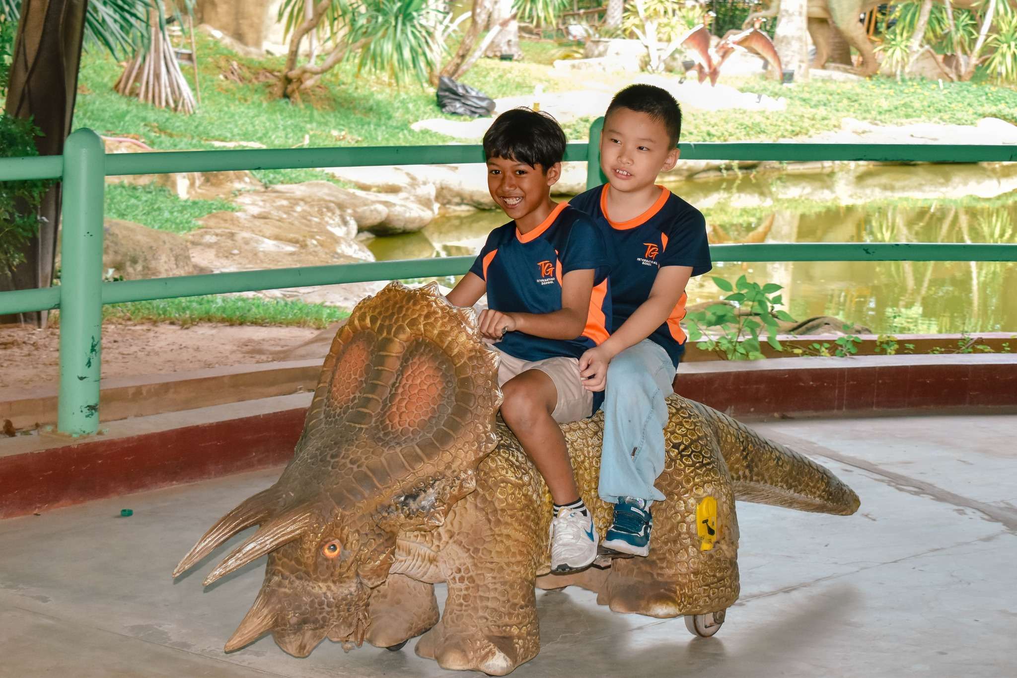 Collage of TG International School students during their Dinosaur Park field trip, including images of children boarding a school bus, a large group photo in front of the park entrance, and students sitting near dinosaur models