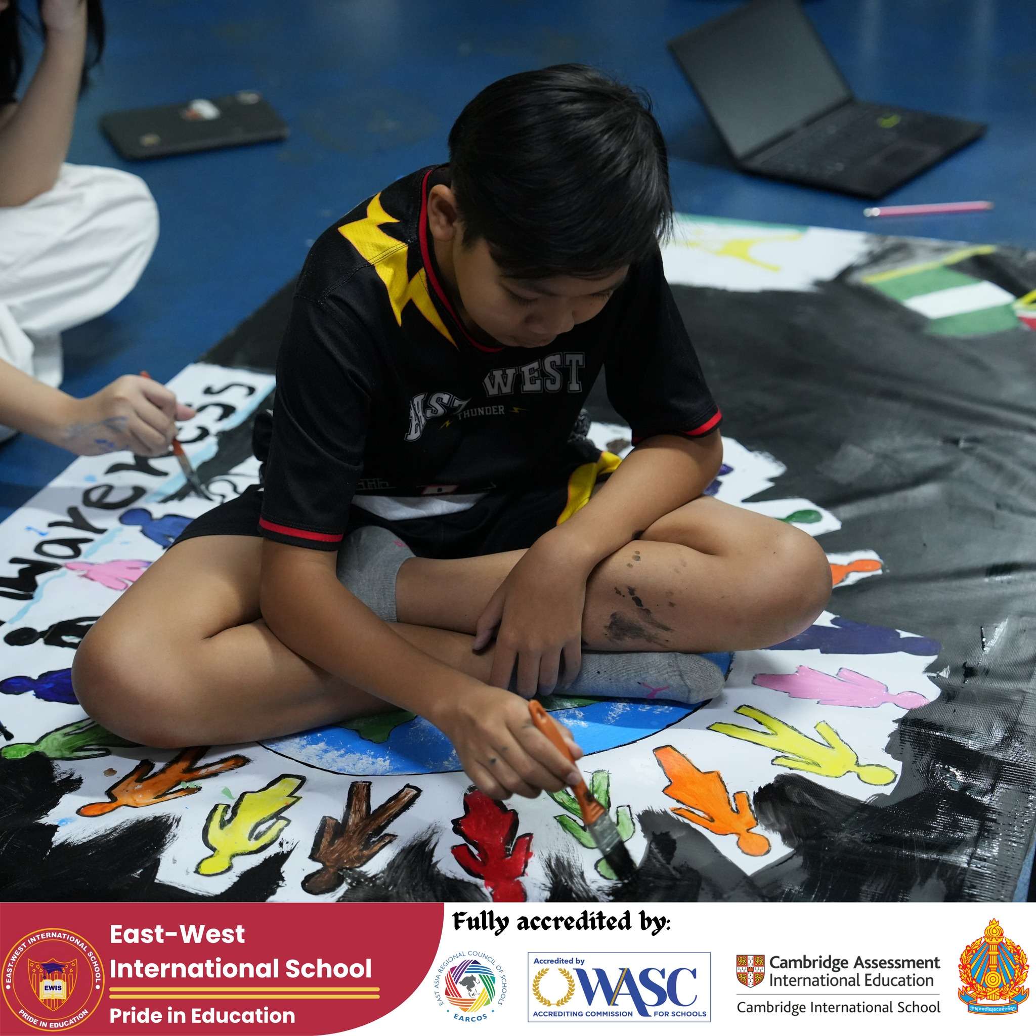 Students from East-West International School Cambodia working together on large-scale painting projects, highlighting creativity and teamwork.