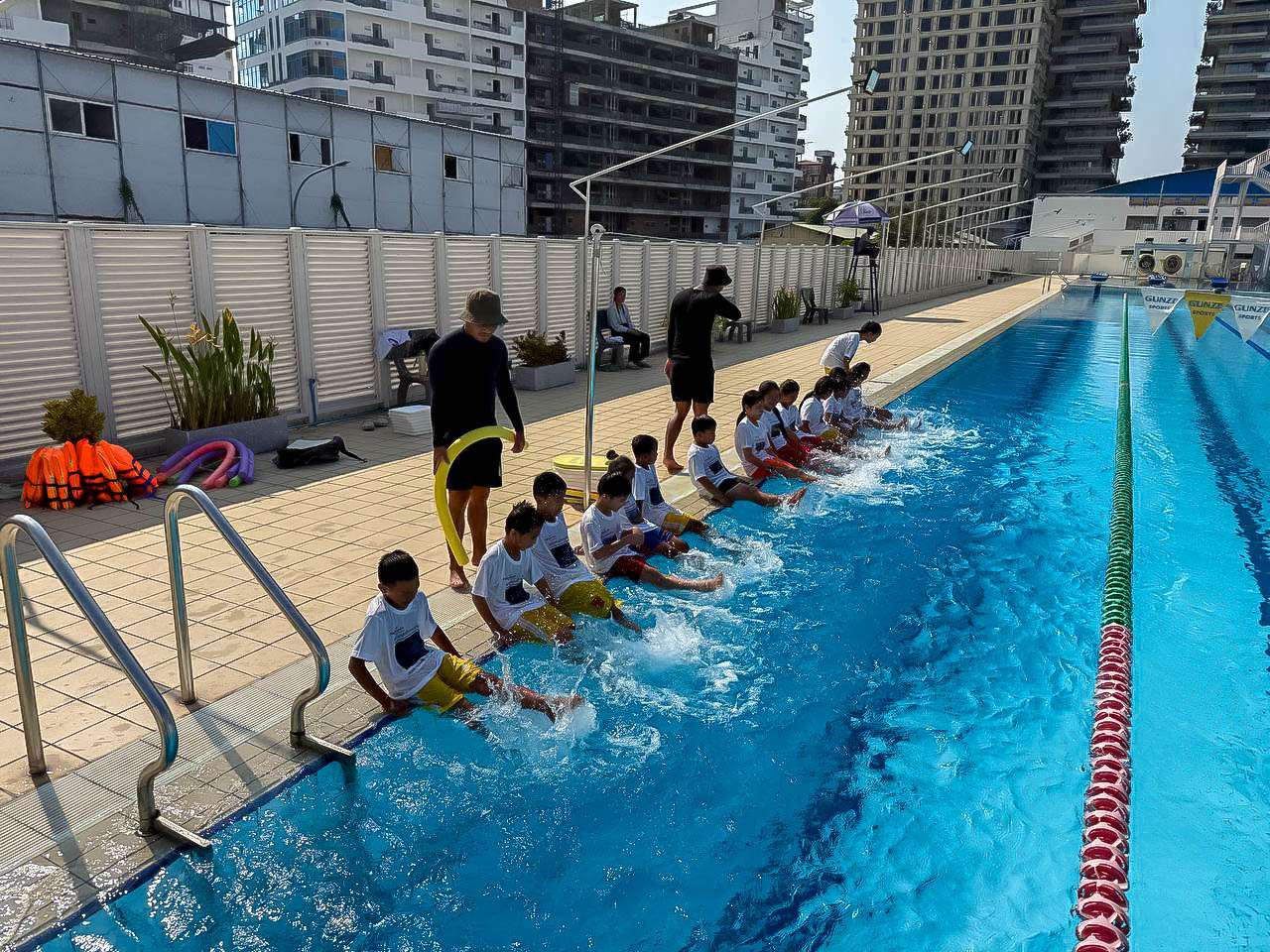 Children practicing warm-up stretches by the pool at Khmer Swimming Federation, swimming lessons at an outdoor pool in Phnom Penh, coaches guiding children in swimming safety and training, group of students learning water skills in a structured program.