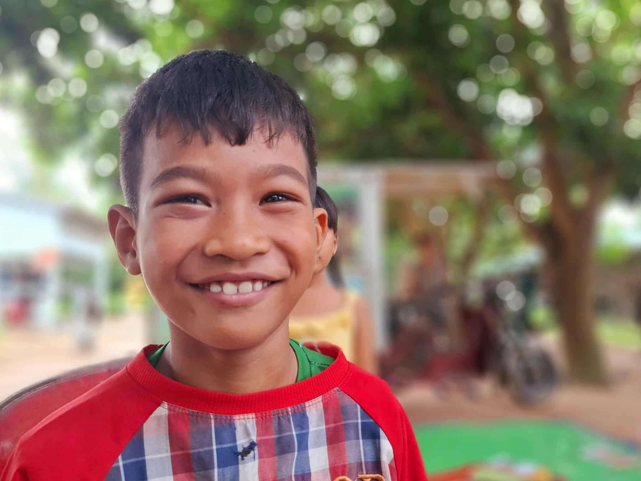 A young Cambodian boy smiles brightly as he stands outdoors, representing the impact of Aide et Action Cambodia’s mobile library initiative bringing books and learning opportunities to rural children.