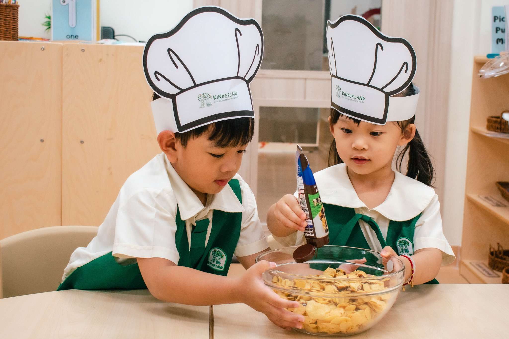 Children from TG International School wearing chef hats and aprons participate in a hands-on cooking activity, mixing ingredients and preparing food as part of a family-themed learning session.
