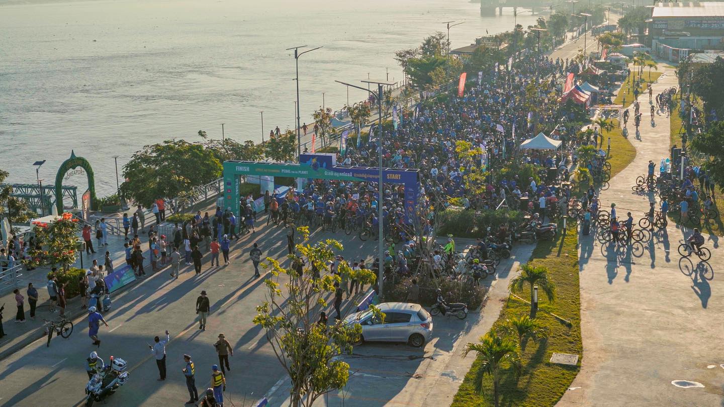 A large community cycling event in Cambodia with participants wearing blue shirts, organized by the European Union promoting environmental awareness.